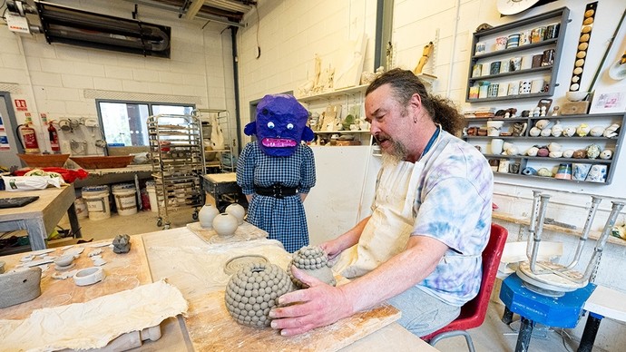 A man with a beard and ponytail and wearing an apron sits in a workshop and holds one hand around an unfired pot made of clay balls; a woman wearing a purple papier-mâché head looks on and listens. 
