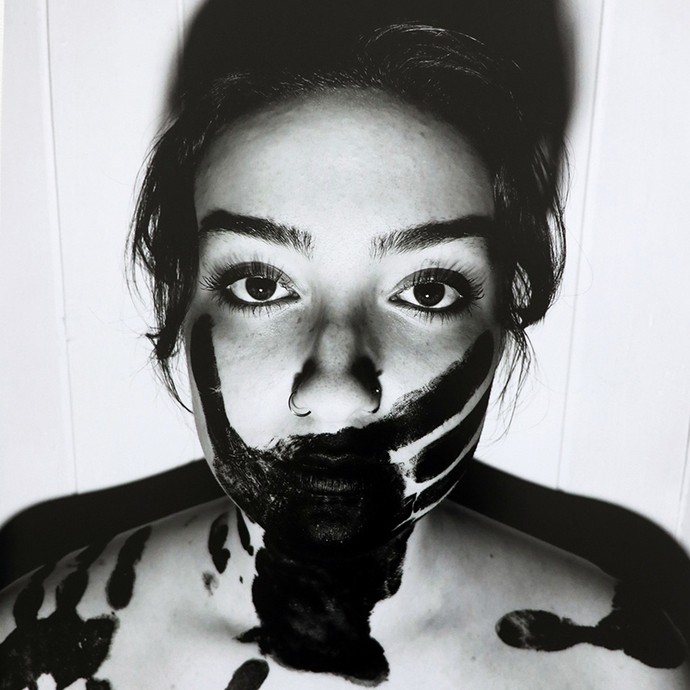 Black-and-white photograph of a young woman with dark hair looking directly into camera; a black handprint covers her mouth; two more black handprints can be seen on her shoulders.