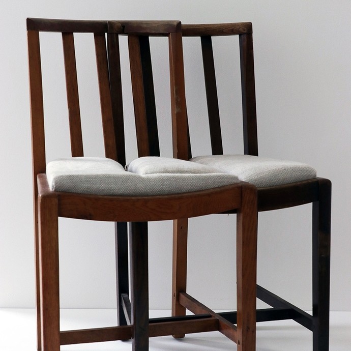 Two wooden dining room chairs merged together; the cross-supports on the chair legs run through each other. 