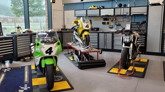 Three motorbikes, including two in racing colours, stand in a workshop with large windows. 