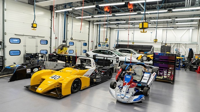 A large well-lit garage with three racing cars in the foreground at different stages of assembly.
