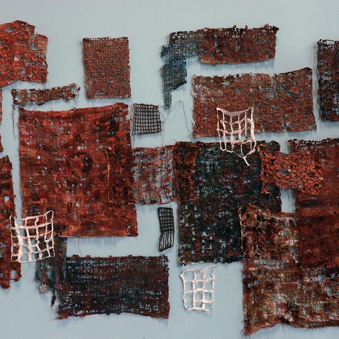 Squares and rectangles of flaking reddish checked cloth in different sizes hang from a wall. 