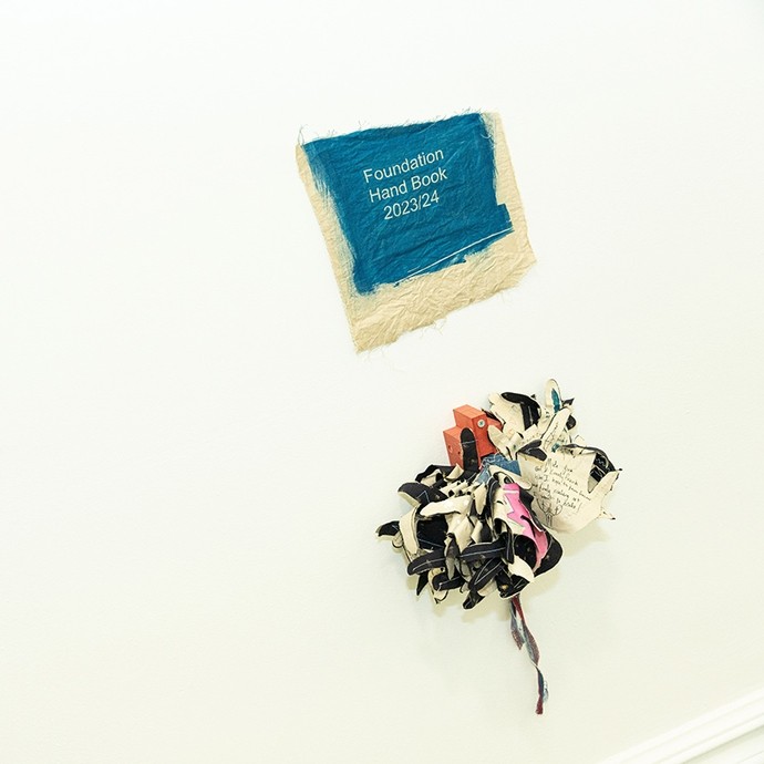 Three objects made of paper on display on a wall; one has a blue cover and reads Foundation Handbook 2023/2024.