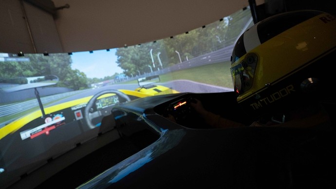 A large screen showing a digital racetrack and car interior; a student wearing a helmet sits in front of the screen with his hands on imitation steering controls.