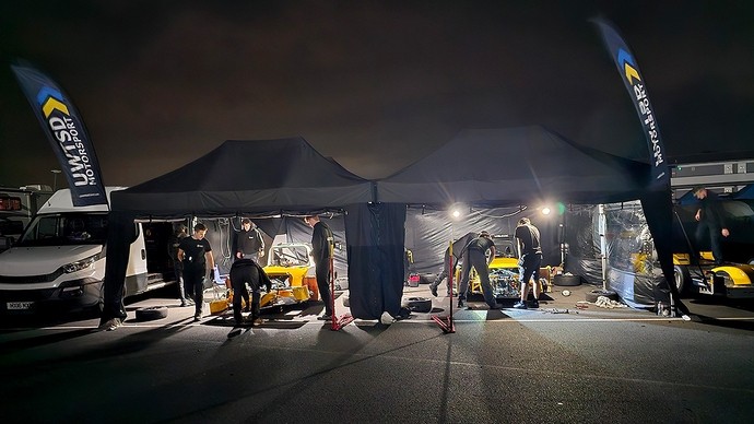 Students work at night on a pair of racing cars in a parking lot; each car is sheltered under a pop-up black gazebo. 