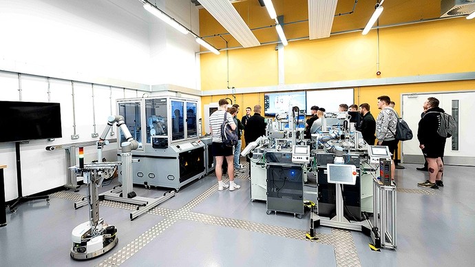 A group of young men inspect the robotics lab; equipment includes a robotic arm and a cyber-physical learning and research platform. 