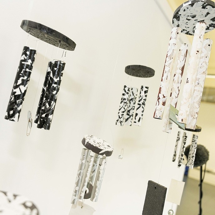 Five sets of windchimes suspended from the ceiling in a white room; each chime is covered in a cross-hatched pattern in black or white, and the top of each set appears to have been recycled or repurposed.