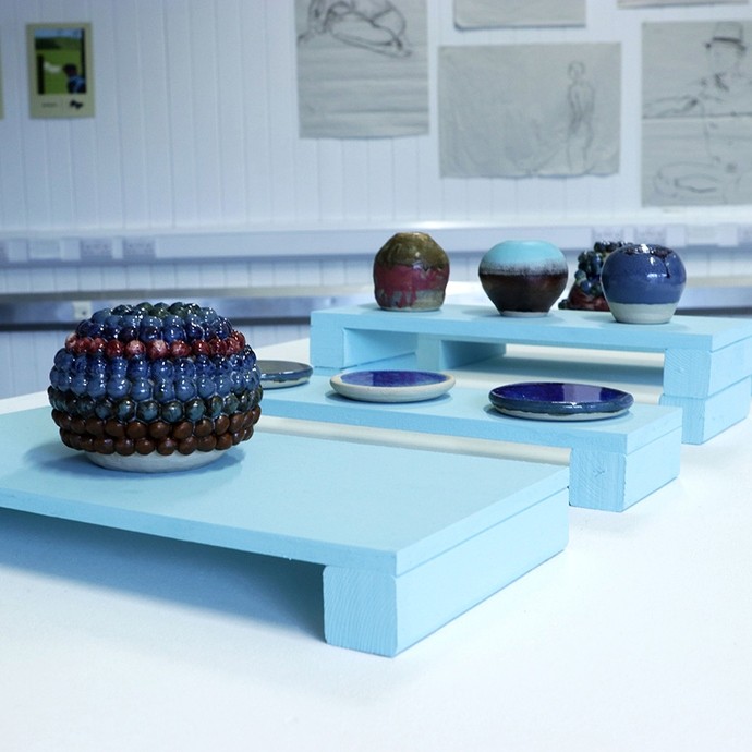 Eight items of ceramic work on display; the nearest is globular and the surface is decorated with nine rows of marble-sized balls; the rows range in colour from brown to blue to red to green.