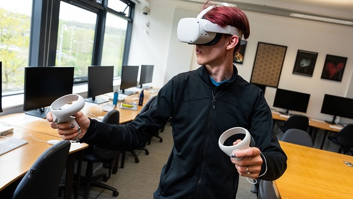 A student stands in a classroom wearing an Oculus Quest headset and holding two controllers.