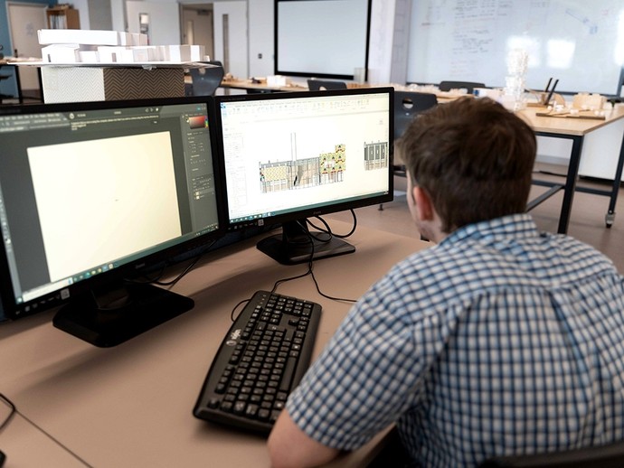 A student sits working in front of dual monitors; one monitor shows Adobe Photoshop while the other shows a building plan. 