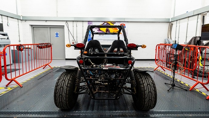 A large go-kart type vehicle with big tyres; it has two seats and an engine at the back exposed to view under the metal frame. 