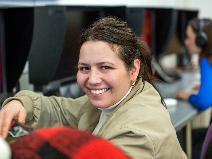 Sitting in front of a computer monitor, a women smiles aside to the camera. 
