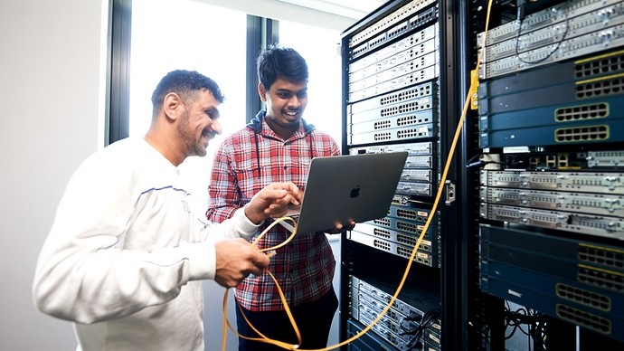 Two smiling men stand next to a server; one holds a MacBook; the other holds a yellow cable attached to the server.