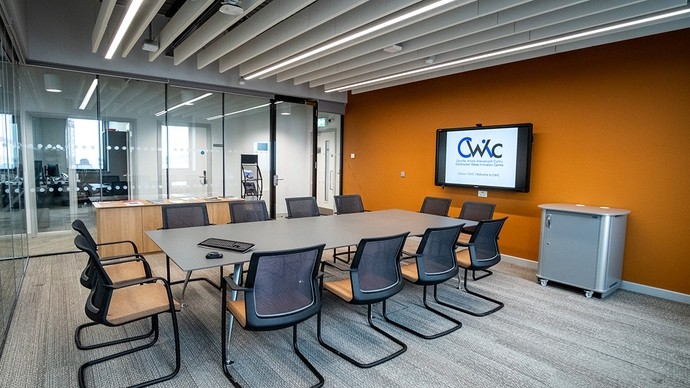 A meeting room with a glass wall on one side; the room is furnished with a large screen TV, a table and chairs.