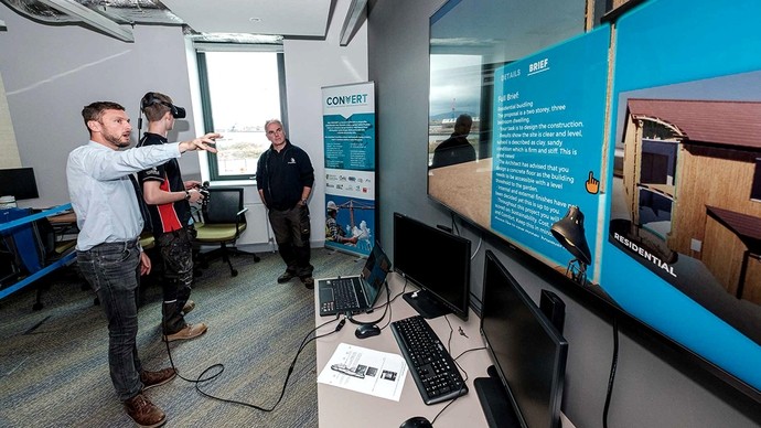 A lecturer gestures towards a screen displaying a 3D building and the word Residential; a student wearing a VR headset listens.