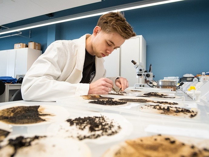A young man in a labcoat inspects a soil sample using tweezers; a microscope can be seen behind him. 