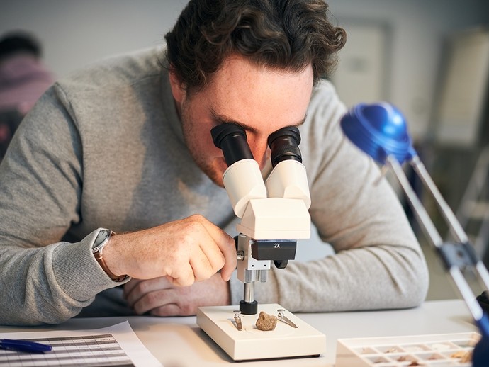 A young man looks through a microscope at a pebble.
