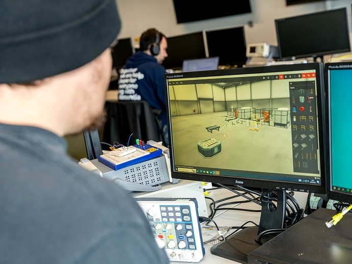 A student looks at a monitor showing a 3D warehouse interior. 