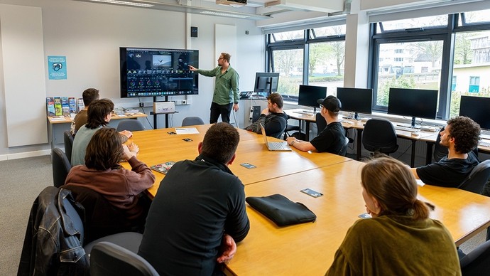 Eight students sit around a table as a lecturer at the head of the room points to a large screen showing a video editing program.