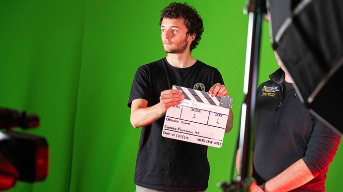 A young man holds a clapperboard and looks off towards the left, waiting; the background is a green screen. 