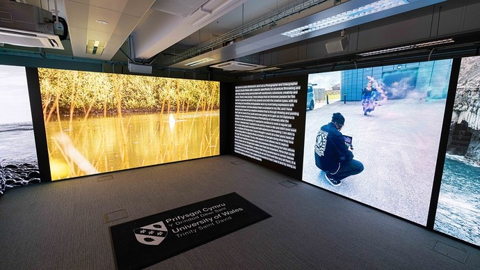 Carmarthen Immersive Room; one wall shows the edge of a lake in sunshine; another shows a woman dancing in a car park amidst coloured smoke.