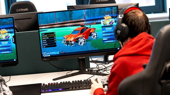 A young man in headphones sits in front of a large monitor displaying a red monster truck.