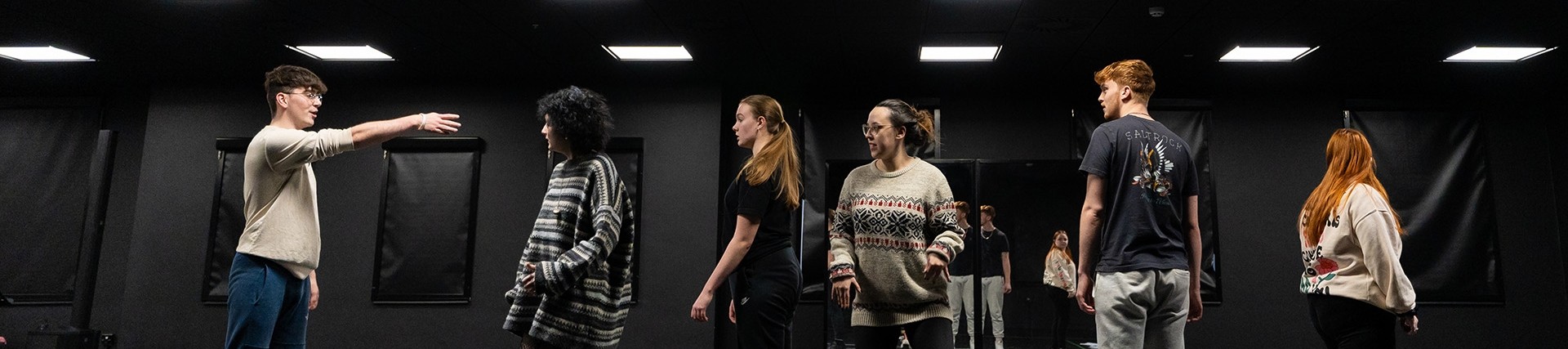 Six students assume different attitudes under the lights of a black-walled rehearsal room. 