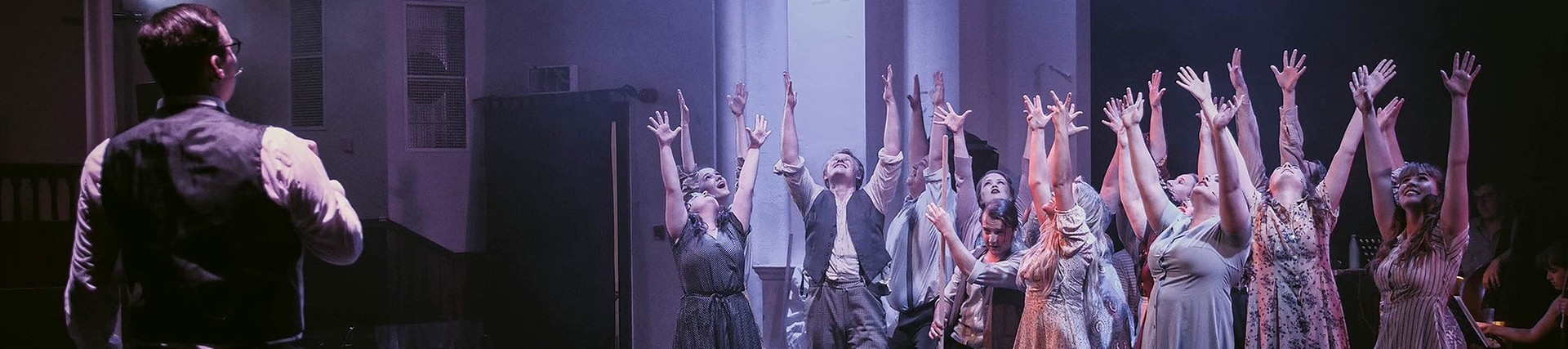 Thirteen cast members dressed in working class early twentieth century costumes raise their hands up as if in joy; a man with parted hair and a black waistcoat watches them from across the stage.