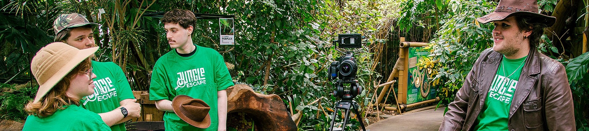 On a path through a tropical glasshouse, a camera stands on a tripod between four young people wearing green Jungle Escape t-shirts.