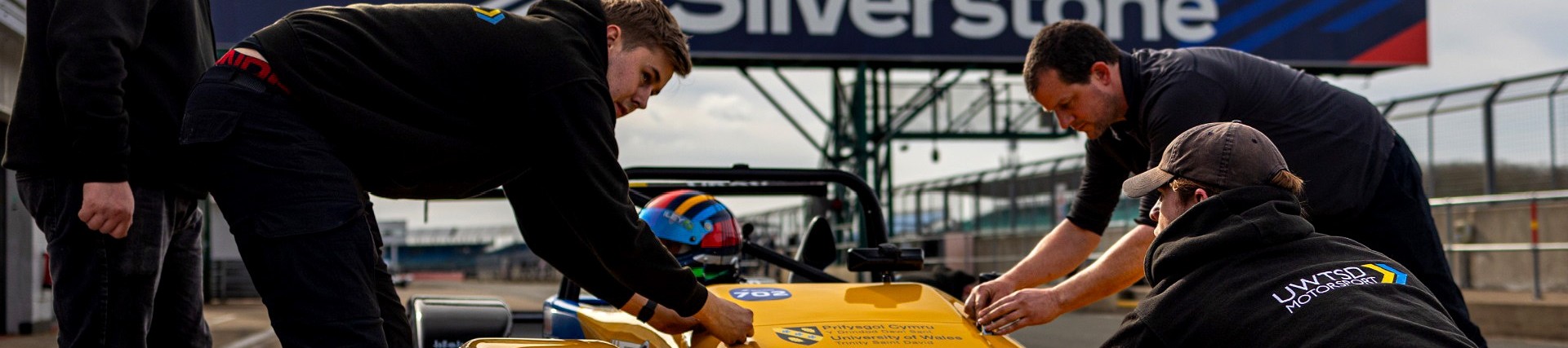 UWTSD Motorsport students bend over a yellow racing car on the track at Silverstone. 