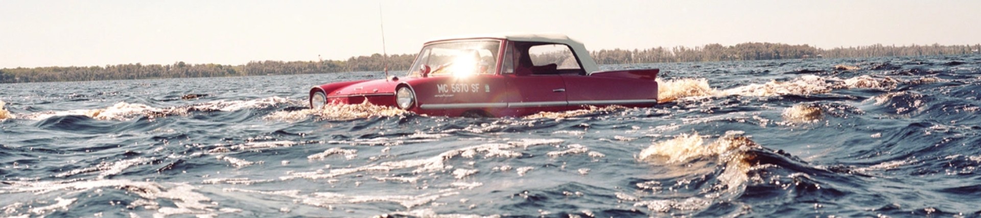 A red sixties cabriolet floats in the middle of a huge lake; the driver looks calm. 