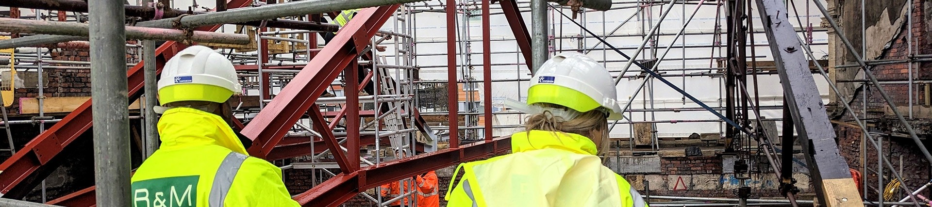 A man and a woman in yellow hi-vis jackets look at scaffolding and supports filling the roof area in the remains of a brick building.