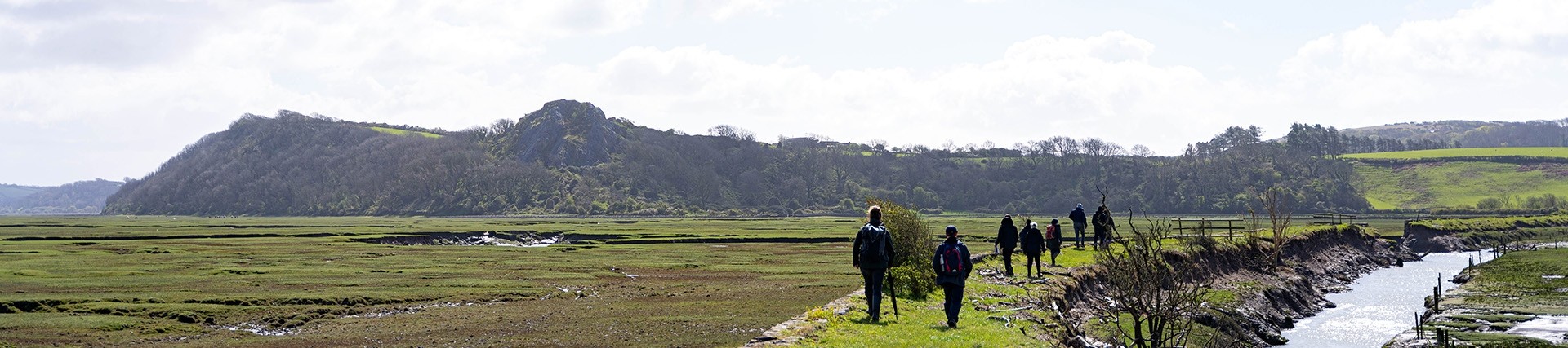 Seven people walk along a dyke with hills in front of them, a stream on their right and an area of tidal marshland on their left.