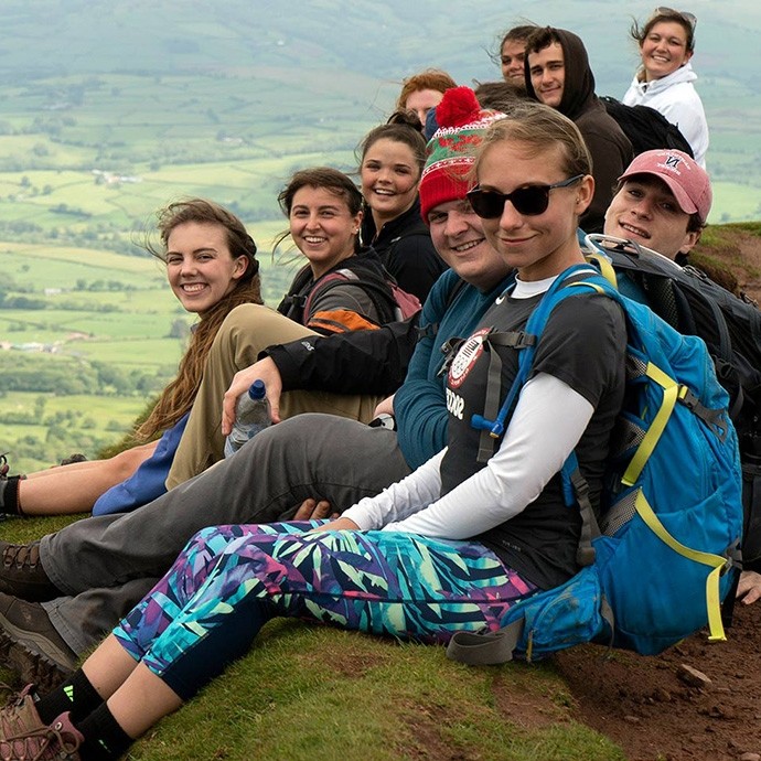 A small group of smiling young people sit on the flank of Pen Y Fan, the green fields of Powys far below them.