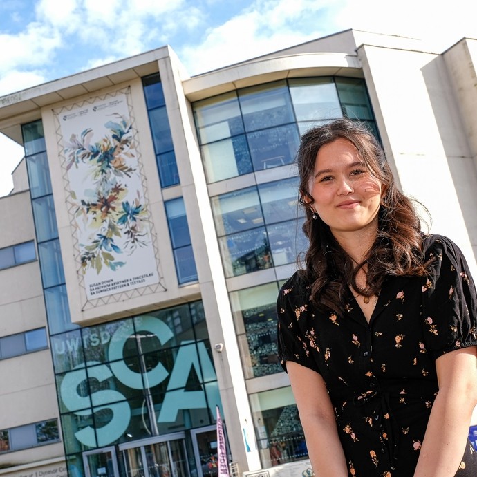 A smiling student standing in front of a giant canvas on the top of a University building in Swansea which features her work.