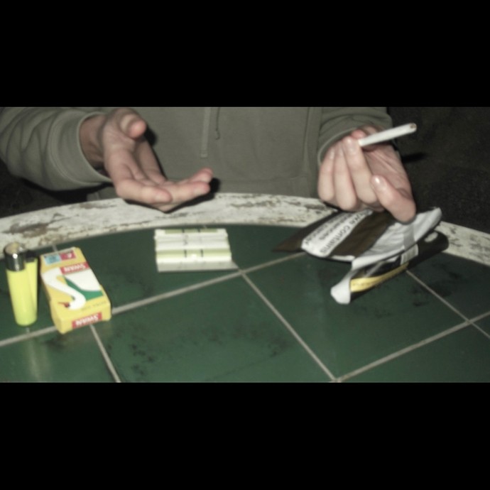 A photograph showing cigarette rolling equipment on a grimy green table and two hands, one holding a cigarette, raised in a gesture that might be invitation. 
