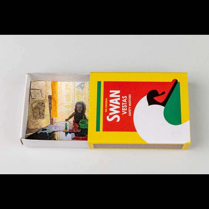 A Swan Vestas Safety Matches box open to reveal underneath its cover part of a painting showing a girl absorbed in making a painting herself. 